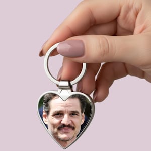 May include: Silver heart-shaped keyring featuring a photograph of a man. The keyring has a silver ring for attaching keys. The photograph is in a heart-shaped frame.
