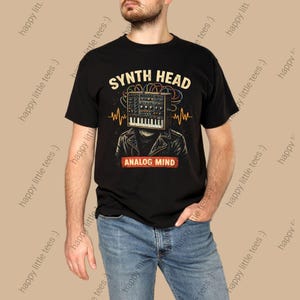 May include: Black t-shirt featuring a graphic of a synthesizer head with the text "SYNTH HEAD" above and "ANALOG MIND" below. The design includes a keyboard, wires, and sound wave lines. The shirt is worn by a person in blue jeans.