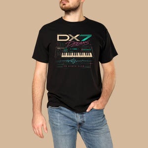 Puede incluir: Camiseta negra con un gráfico de teclado de sintetizador y el texto "DX7 Dreams" y "FM Synth Club" en verde azulado y rosa. La camiseta tiene cuello redondo y mangas cortas.