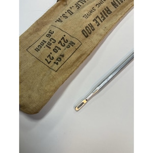 May include: A vintage rifle cleaning rod with a canvas storage sleeve. The sleeve is tan with black text that reads "IN RIFLE ROD" and "36 inch". The rod is silver and has a loop at the end. The text also reads "No. 101 .22 to .27 Cal." and "CALIF., U.S.A."