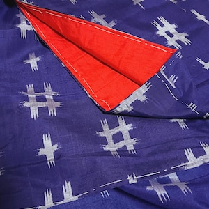 Puede incluir: Un textil doblado con un interior rojo vibrante y un exterior azul oscuro. La tela azul está estampada con formas blancas similares a hashtags. Los bordes están cuidadosamente cosidos con hilo blanco. La estética general es moderna y gráfica.