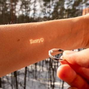 May include: A silver ring held in a hand, with the word "Henry" and a heart symbol projected onto the arm. The image is set against a blurred outdoor background, with the text "Of Elven Make" at the bottom.