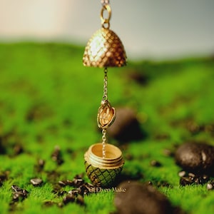 Puede incluir: Un colgante dorado y ornamentado con una parte superior en forma de campana y una pequeña parte inferior abierta. El colgante cuelga de una delicada cadena, con un pequeño dije de forma ovalada en el medio. El colgante está sobre un fondo de musgo verde y pequeñas piedras negras.