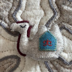 Op de afbeelding: Een vilten kameelfiguur in lichtgrijs met bordeauxrode accenten. De kameel heeft een blauwe patch met een rood ontwerp en gele stiksels. De kameel staat op een getextureerde, gebroken witte achtergrond met grijze stiksels.