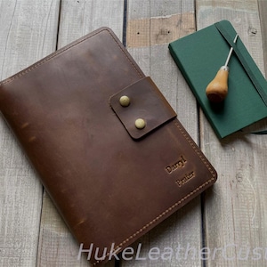 May include: A brown leather journal with a flap closure and brass-coloured studs. The journal is personalised with the name "Darryl Prater". A green notebook and a wooden and metal tool are next to the journal. The journal and notebook are on a wooden surface.