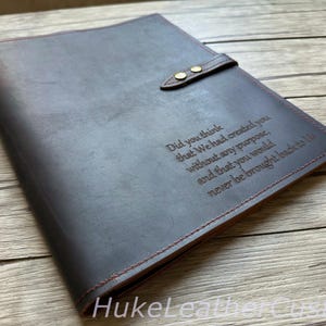 May include: Dark gray leather journal with a snap closure and red stitching. The cover features engraved text: "Did you think that We had created you without any purpose, and that you would never be brought back to Us."