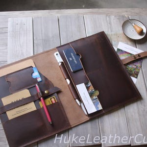 Könnte beinhalten: Geöffnetes braunes Lederportfolio mit mehreren Taschen für Karten, einen Stift und ein Telefon. Das Portfolio ist offen und zeigt einen hellbraunen Innenraum. Ein roter Stift und eine blaue Karte sind sichtbar. Das Portfolio liegt auf einer Holzoberfläche.