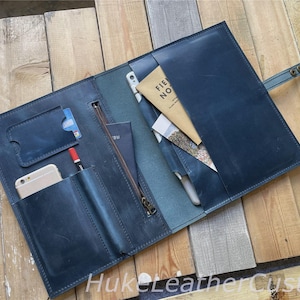 Può includere: Cartella in pelle blu scuro aperta, con più tasche, una cerniera e una chiusura a scatto. La cartella contiene uno smartphone, una penna, carte e un taccuino "Field Notes". La cartella è aperta su una superficie di legno.