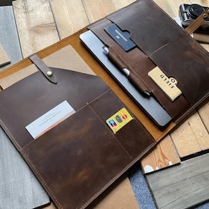 Puede incluir: Un portafolio de cuero marrón con múltiples bolsillos, que contiene una tableta, un bolígrafo y tarjetas. El portafolio está abierto, revelando un bloc de notas y una pequeña etiqueta rectangular que dice "FIELD". Una cámara vintage descansa cerca.