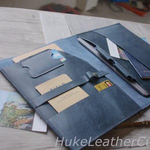 Puede incluir: Un folio de cuero azul con múltiples bolsillos y ranuras para tarjetas. El folio contiene una tableta, un bolígrafo y varias tarjetas. El folio está abierto, revelando el interior. El texto "HukeLeatherCustom" es visible.