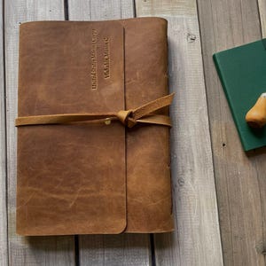 May include: A brown leather journal with a strap closure and the text "United States Marine Corps, Vlahakis, Mason C." A green notebook and a leather-handled awl are next to the journal.