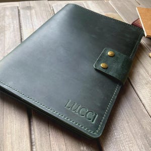 May include: Dark green leather journal with the embossed word "LUCCI" on the cover. The journal has a strap closure with two brass-coloured snaps. Additional notebooks are visible in the background.