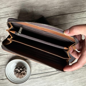 May include: Open brown leather wallet with multiple compartments and a zip closure. The wallet is held open, revealing the interior. A small, decorative white bowl with a pinecone is in the lower left corner.