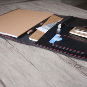 Puede incluir: Portafolio de cuero negro con costuras rojas, abierto para revelar un bloc de notas, un teléfono inteligente y un cuaderno pequeño. Una tarjeta de crédito es visible en un bolsillo. El portafolio está sobre una superficie de madera.