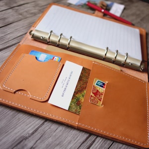 Puede incluir: Carpeta de cuero marrón claro abierta, con anillas plateadas, que contiene papel rayado. La carpeta tiene ranuras para tarjetas y un bolsillo pequeño. Un bolígrafo rojo descansa sobre la superficie de madera.