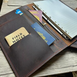 Może przedstawiać: Brązowe skórzane portfolio z kieszenią zapinaną na zamek, mieszczące paszport, książeczkę Field Notes i niebieską kartę. Portfolio jest otwarte, ukazując notatnik z liniowanym papierem i długopisem. Widoczny jest tekst "FIELD NOTES".