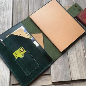 以下が含まれることがあります： 開いた濃い緑色の革製ノートカバー。中にはベージュ色のノートが入っています。カバーにはカードやその他のアイテム用のポケットがあります。小さな赤い本とポストカードが、木製の表面にあるノートの隣にあります。