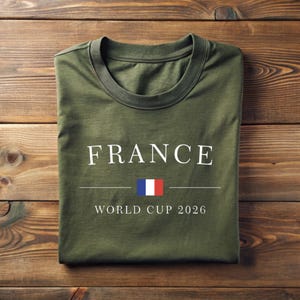 May include: Olive green t-shirt with the word "FRANCE" in white capital letters, a horizontal line, and the French flag. Below the line, the text reads "WORLD CUP 2026". The shirt is folded on a wooden surface.