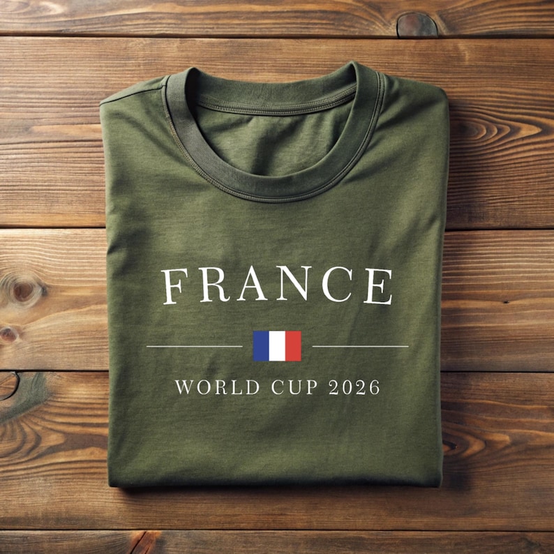 May include: Olive green t-shirt with the word "FRANCE" in white capital letters, a horizontal line, and the French flag. Below the line, the text reads "WORLD CUP 2026". The shirt is folded on a wooden surface.