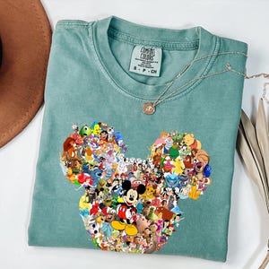 May include: A seafoam green t-shirt with a Mickey Mouse head design made up of various Disney characters. The shirt has a crew neck and short sleeves. A silver necklace with a small pendant rests on the shirt.