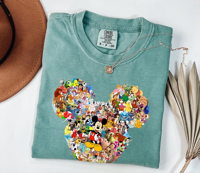 May include: A seafoam green t-shirt with a Mickey Mouse head design made up of various Disney characters. The shirt has a crew neck and short sleeves. A silver necklace with a small pendant rests on the shirt.