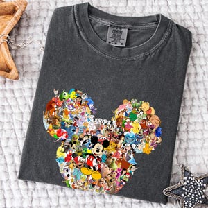 May include: Dark grey t-shirt with a Mickey Mouse head design made up of various Disney characters. The shirt has a crew neck and is made of a soft material. The brand name "Comfort Colors" is visible on the tag.