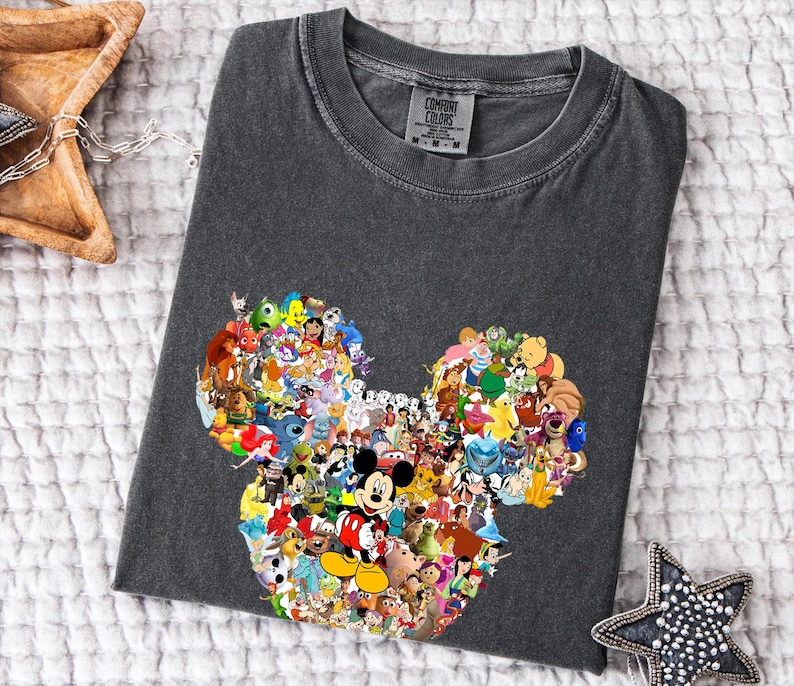 May include: Dark grey t-shirt with a Mickey Mouse head design made up of various Disney characters. The shirt has a crew neck and is made of a soft material. The brand name "Comfort Colors" is visible on the tag.
