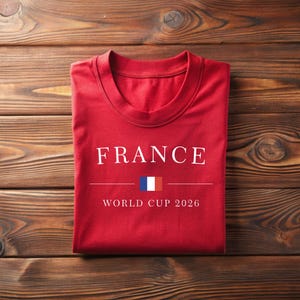 May include: A folded red t-shirt with the word "FRANCE" in white capital letters. Below the word is a horizontal line, a small French flag, and the text "WORLD CUP 2026". The shirt is on a wooden surface.