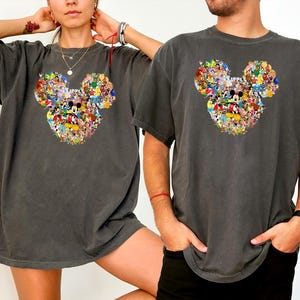 May include: Two charcoal grey t-shirts with a Mickey Mouse head design filled with cartoon characters. The shirts have short sleeves and a relaxed fit, suitable for casual wear. The design is colourful and eye-catching.