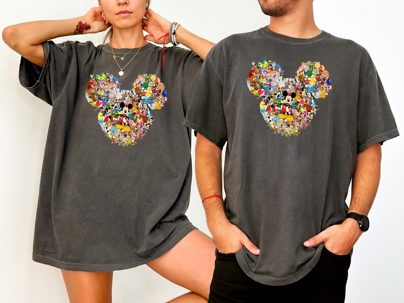 May include: Two charcoal grey t-shirts with a Mickey Mouse head design filled with cartoon characters. The shirts have short sleeves and a relaxed fit, suitable for casual wear. The design is colourful and eye-catching.