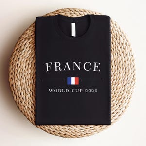 May include: Black t-shirt with the word "FRANCE" in white capital letters. Below the word is a horizontal line, the French flag, and the text "WORLD CUP 2026". The shirt is displayed on a woven, circular surface.