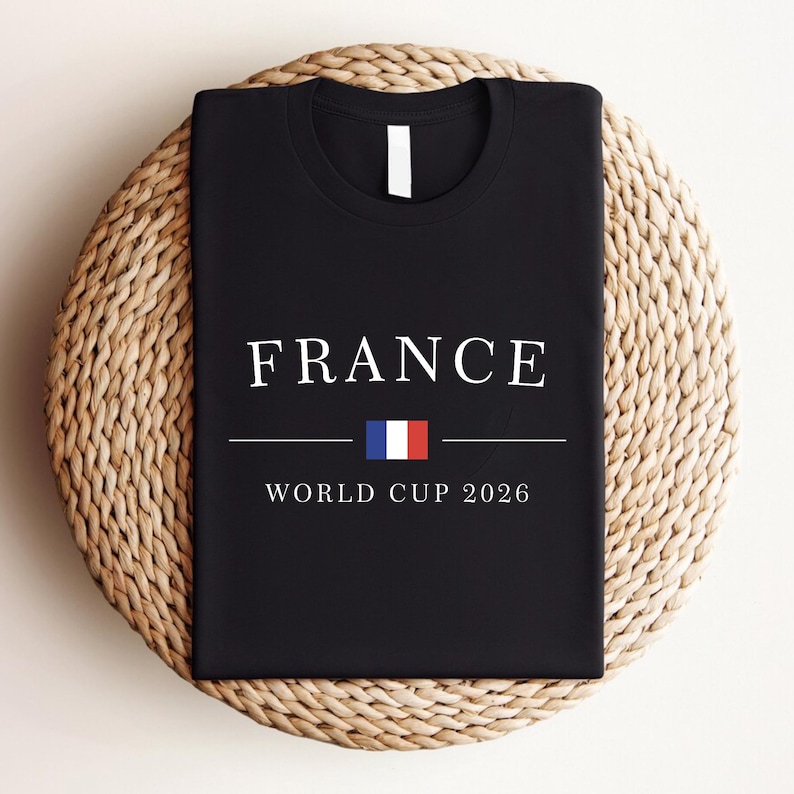 May include: Black t-shirt with the word "FRANCE" in white capital letters. Below the word is a horizontal line, the French flag, and the text "WORLD CUP 2026". The shirt is displayed on a woven, circular surface.