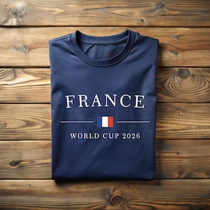 May include: A navy blue t-shirt with the word "FRANCE" in white capital letters. Below the word is a horizontal line, a small French flag, and the text "WORLD CUP 2026". The shirt is folded neatly on a wooden surface.