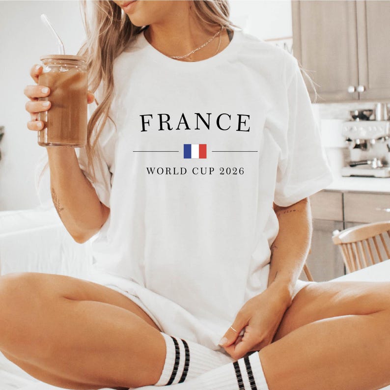 May include: White t-shirt with the word "FRANCE" in black capital letters, a horizontal line, and the text "WORLD CUP 2026" below. The French flag is between the line and the text. The person is wearing black and white striped socks.