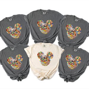 May include: Six t-shirts in shades of grey and cream. Each shirt displays a Mickey Mouse head design, composed of various colourful cartoon characters. The shirts are made of a soft material with short sleeves.