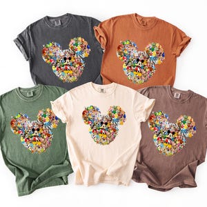May include: Five t-shirts in shades of grey, orange, green, cream, and brown. Each shirt displays a Mickey Mouse head design composed of Disney character images. The t-shirts are short-sleeved and have a relaxed fit.