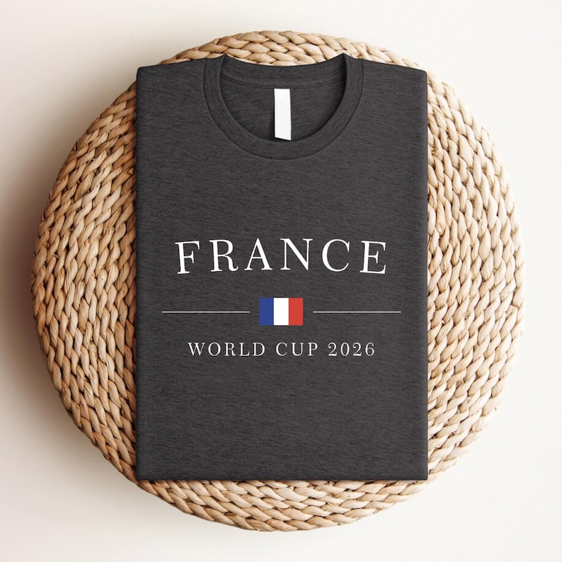 May include: Dark gray t-shirt with the word "FRANCE" in white capital letters. Below the word is a horizontal line, a small French flag, and the text "WORLD CUP 2026". The shirt is displayed on a round, woven wicker placemat.