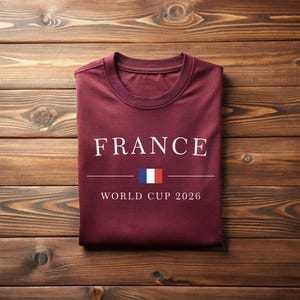 May include: A maroon t-shirt with the word "FRANCE" in white capital letters. Below the word is a horizontal line with the French flag and the text "WORLD CUP 2026". The shirt is folded and lies on a wooden surface.