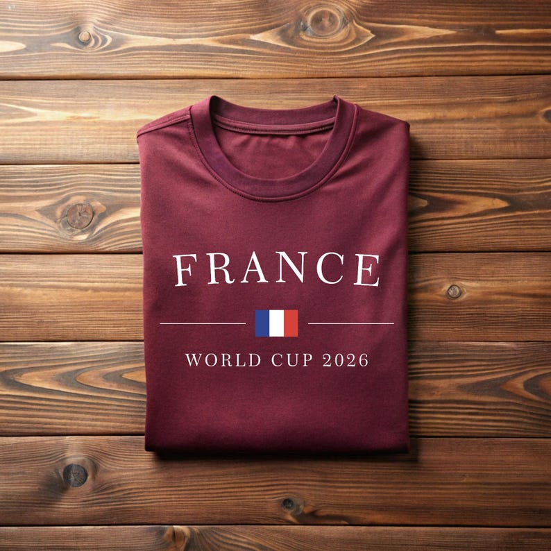 May include: A maroon t-shirt with the word "FRANCE" in white capital letters. Below the word is a horizontal line with the French flag and the text "WORLD CUP 2026". The shirt is folded and lies on a wooden surface.
