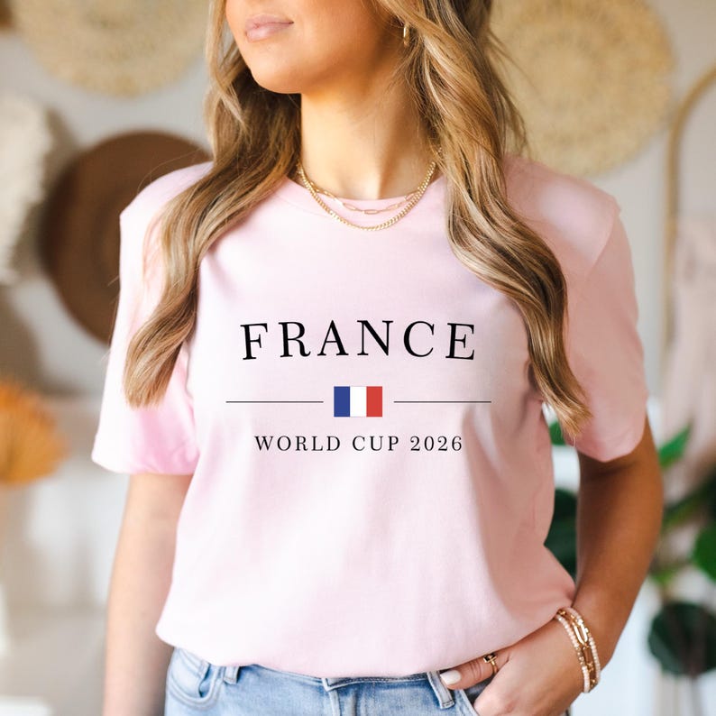 May include: A light pink t-shirt with the word "FRANCE" in black capital letters, a horizontal line, and the French flag in blue and red. Below the line, the text reads "WORLD CUP 2026".