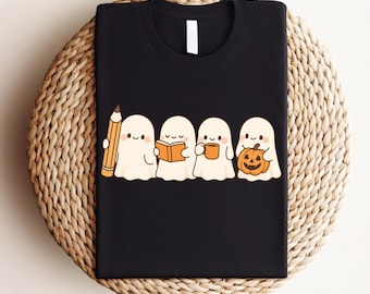May include: Black t-shirt featuring a cute cartoon design of four ghosts. One ghost holds a pencil, another a book, a third a mug, and the last a jack-o'-lantern. The ghosts are white with small black eyes and rosy cheeks.