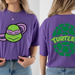 May include: Purple t-shirt featuring a green and purple cartoon character on the front. The back of the shirt displays a green turtle shell design with the word "TURTLES" in green. The shirt is made of a soft material.
