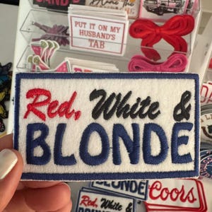 May include: A white rectangular patch with the words "Red, White & BLONDE" embroidered in red, black, and blue. Other patches with text and designs are visible in the background, including "Coors" and "BRONCO".