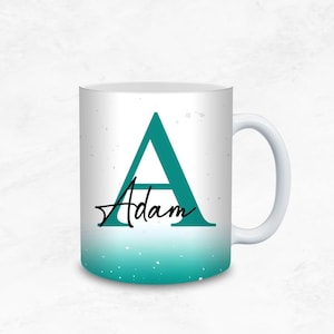 Pode incluir: Caneca de cerâmica branca com um gradiente turquesa na parte inferior e uma grande letra "A" turquesa. O nome "Adam" está escrito em itálico abaixo da letra. A caneca tem uma alça branca e um fundo branco.