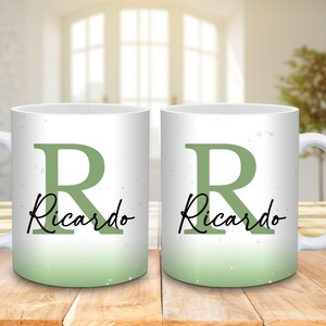 May include: Two white ceramic mugs with a green ombre design. Each mug features a large green letter "R" and the name "Ricardo" in a black script font. The mugs are on a wooden surface.