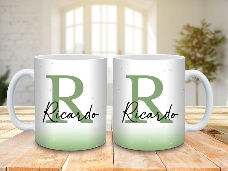 May include: Two white ceramic mugs with a green ombre design. Each mug features a large green letter "R" and the name "Ricardo" in a black script font. The mugs are on a wooden surface.