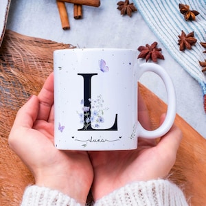 Könnte beinhalten: Weiße Keramiktasse mit einem großen schwarzen Buchstaben "L", verziert mit lila Blumen und Schmetterlingen. Der Name "Luna" steht darunter. Die Tasse hat einen gebogenen Henkel und wird von einer Person gehalten.