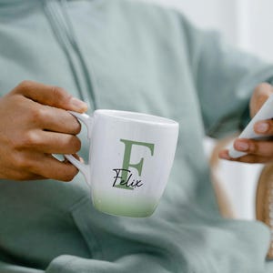 May include: White ceramic mug with a green ombre effect at the base. The mug features a large green letter "F" and the name "Felix" in a script font. The mug is held by a person.