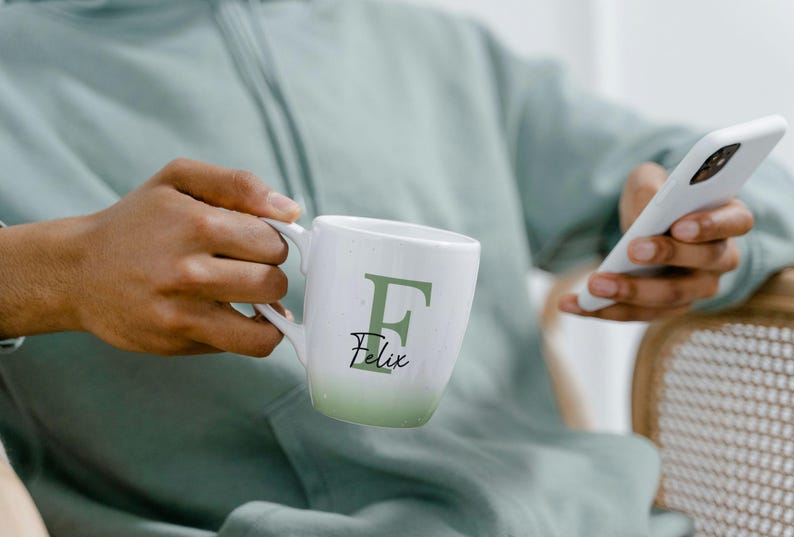 May include: White ceramic mug with a green ombre effect at the base. The mug features a large green letter "F" and the name "Felix" in a script font. The mug is held by a person.