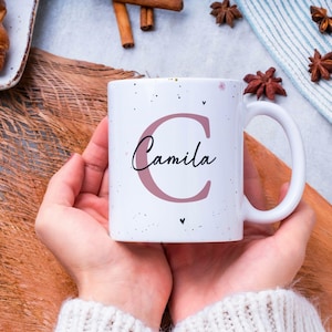 Puede incluir: Una taza de cerámica blanca con el nombre "Camila" en escritura cursiva, con una gran "C" en color malva. La taza se sostiene con ambas manos, con un fondo de pasteles y especias.